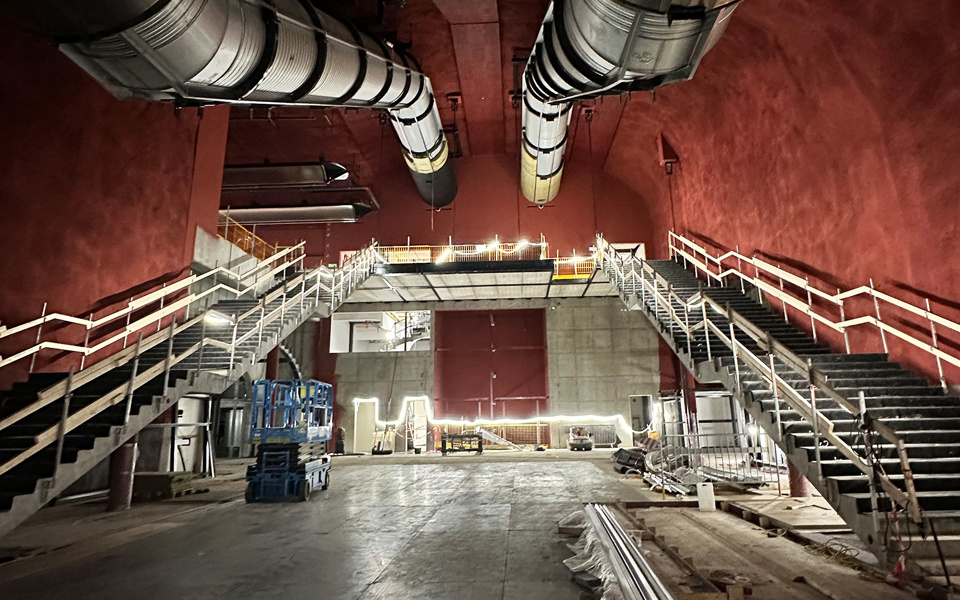 Byggområde inne i ett bergrum. Tak och väggar är rödmålade. På höger och vänstersida leder två trappor upp till ett balkongplan. I taket går två stora ventilationskanaler.